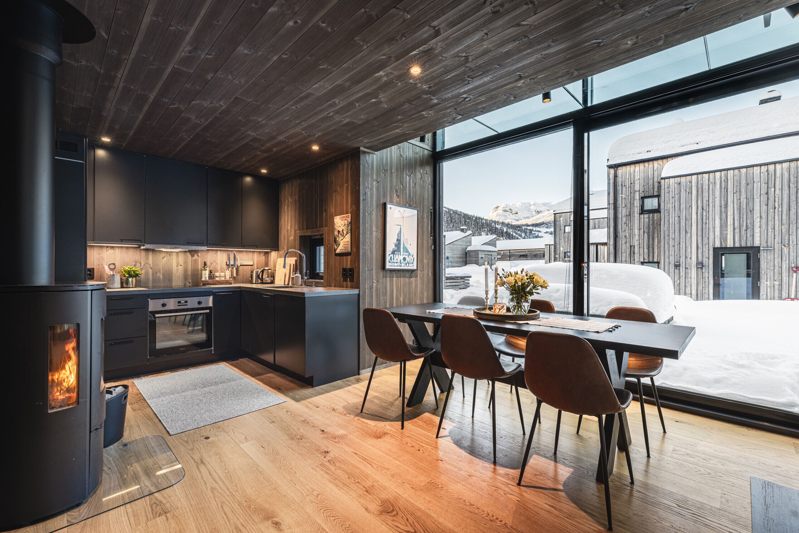 Una cocina moderna con estufa de leña, comedor con sillas y un ventanal con vistas a un paisaje nevado.