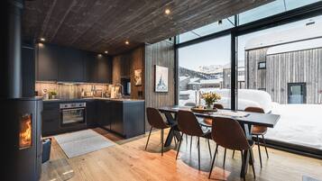 Una cocina moderna con estufa de leña, comedor con sillas y un ventanal con vistas a un paisaje nevado.