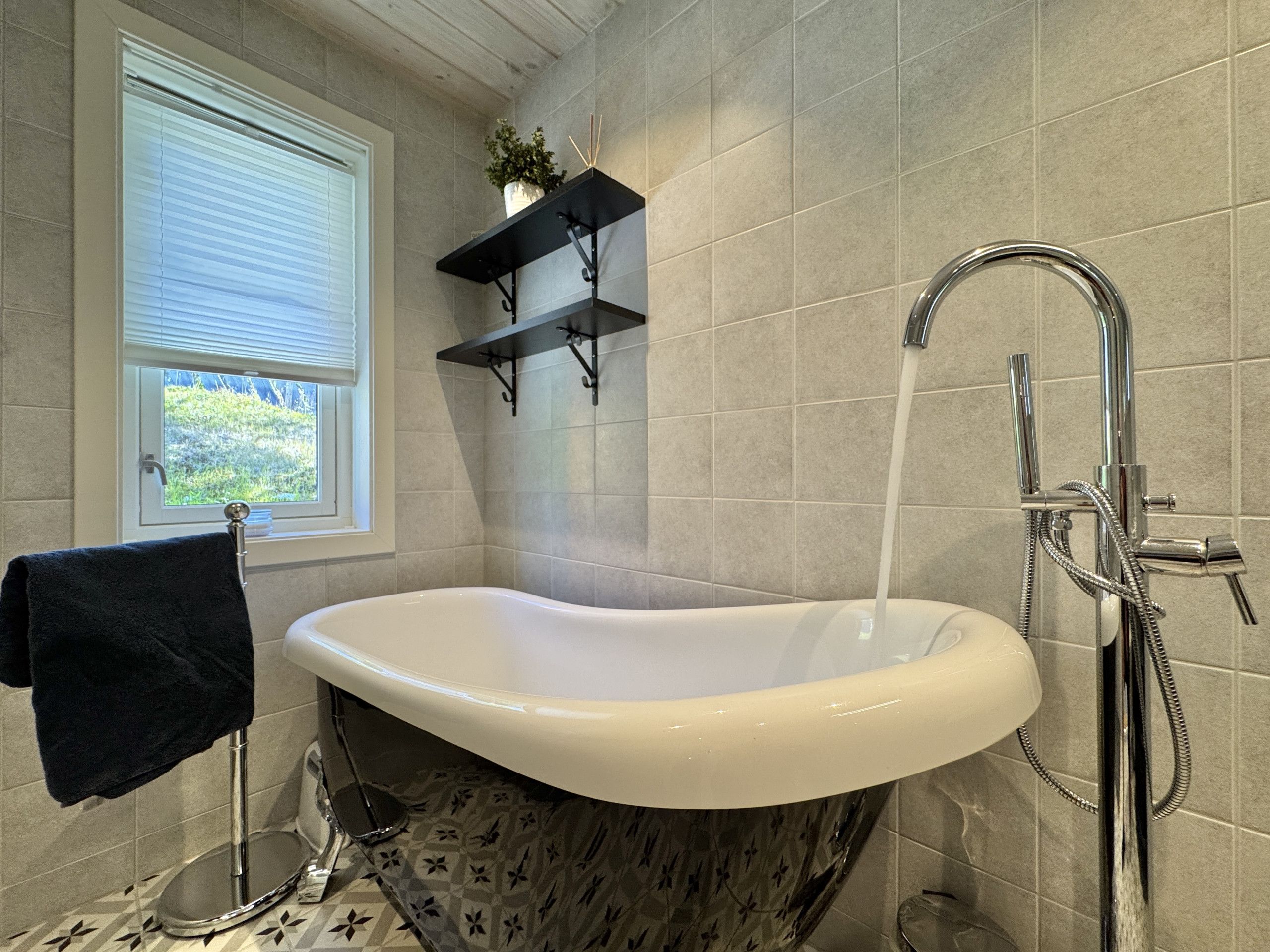 A bathroom with a freestanding bathtub, a window with blinds, a towel rack, and a shower with a handheld showerhead.