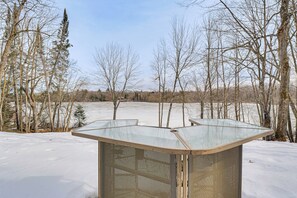 A snow-covered outdoor area with a metal structure, possibly a shelter, surrounded by leafless trees. - Lakefront Tiny Home w/ Dock Near Chetek! (Bruce)