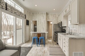 A kitchen with white cabinets, a granite countertop, and a built-in microwave. There are two blue stools and a window overlooking a snowy landscape. - Lakefront Tiny Home w/ Dock Near Chetek! (Bruce)