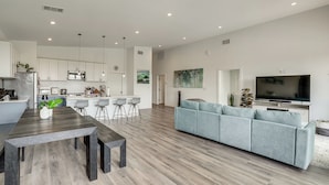 Eine moderne Küche mit weißen Schränken, ein Essbereich mit einem Holztisch und Bänken, ein graues Sofa und ein Flachbildfernseher.