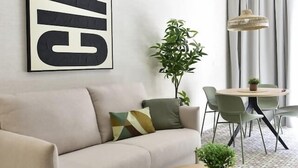 Ein modernes Wohnzimmer mit einer Couch, einem runden Couchtisch, einem großen Wandbild, einem Topf mit Pflanze sowie einem Essbereich mit Stühlen und Tisch.