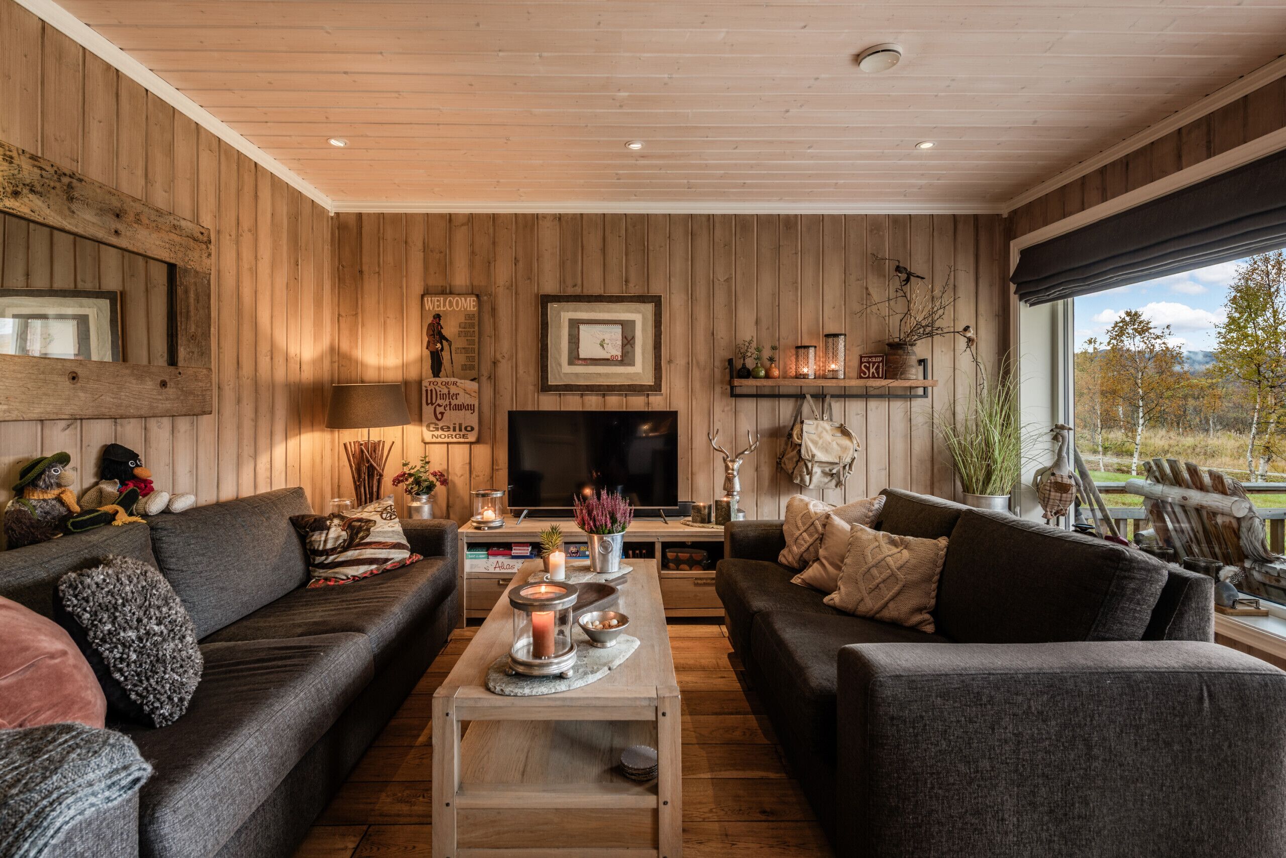 A cozy living room with wooden walls, a large TV, two sofas, a coffee table with candles, and a window with a view.