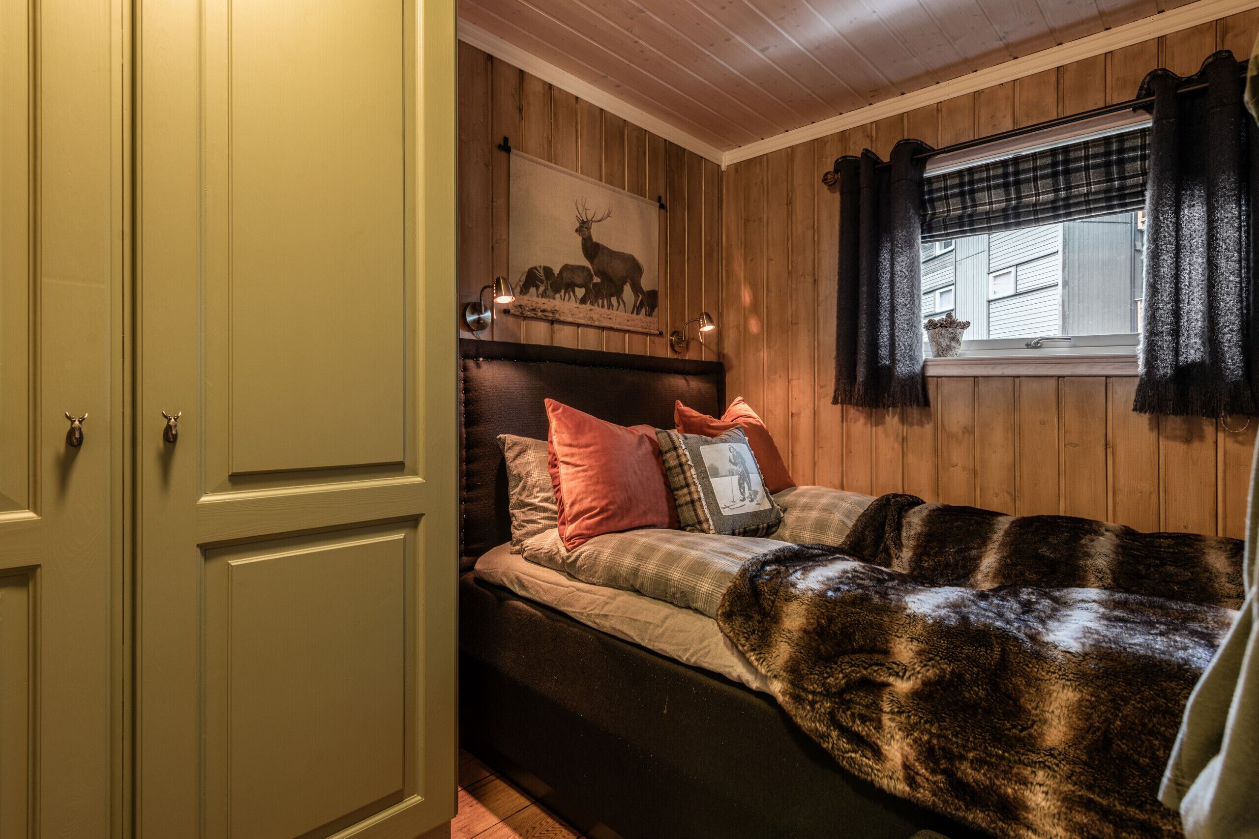 Ein Zimmer mit Holzvertäfelung, einem Bett mit einem Fellüberwurf, einem Wandbild von Hirschen und einem Fenster mit Vorhängen.