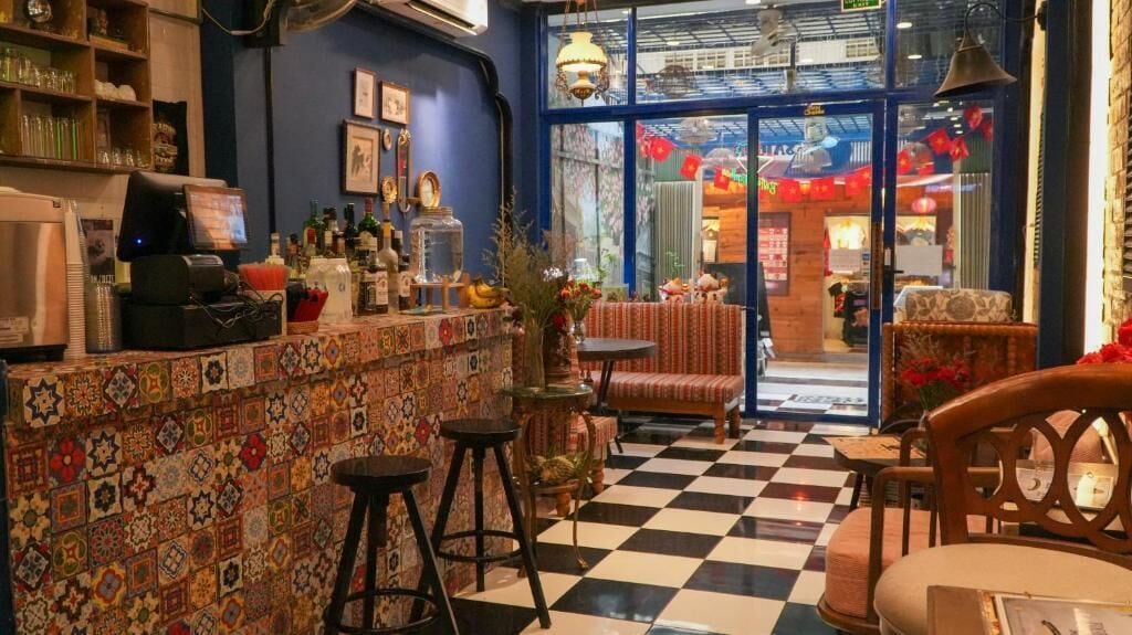 Zona de bar con suelo a cuadros, taburetes y mostrador con diversos objetos.