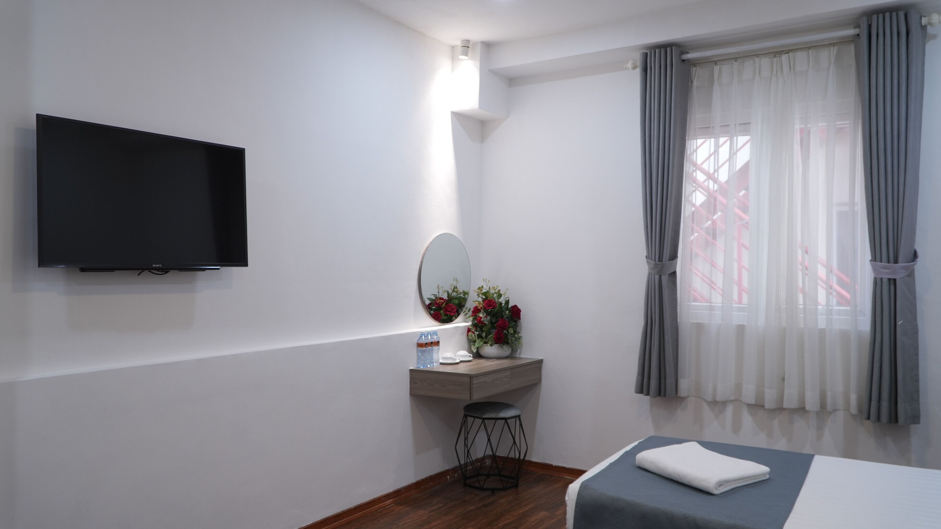 A hotel room with a flat-screen TV, a vanity with a mirror and flowers, a bedside table with a towel, and a window with curtains.