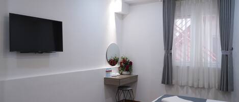 A hotel room with a flat-screen TV, a vanity with a mirror and flowers, a bedside table with a towel, and a window with curtains.