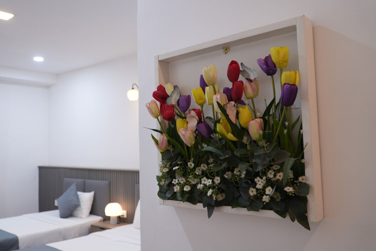 Een hotelkamer met een bed, een nachtkastje met een lamp, en een bloemstuk aan de muur.