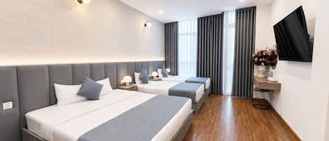 Habitación de hotel con tres camas, un televisor de pantalla plana, una mesita con un florero y un ventanal con cortinas.