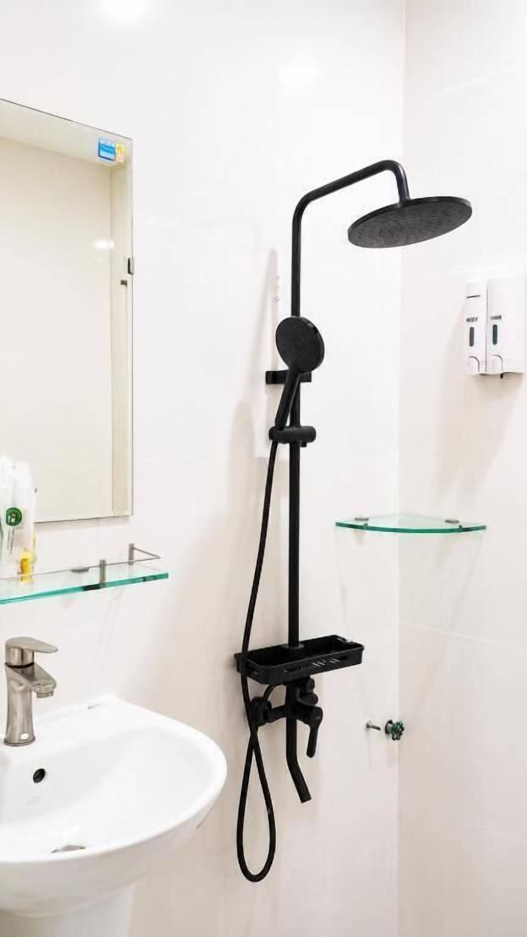Un baño con lavamanos blanco, repisa de vidrio, cabezal de ducha negro y espejo.