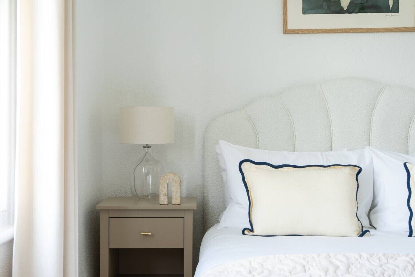 Una cama bien hecha con ropa de cama blanca y almohadas azul marino, una lámpara de noche y una mesita de noche pequeña con un cajón.