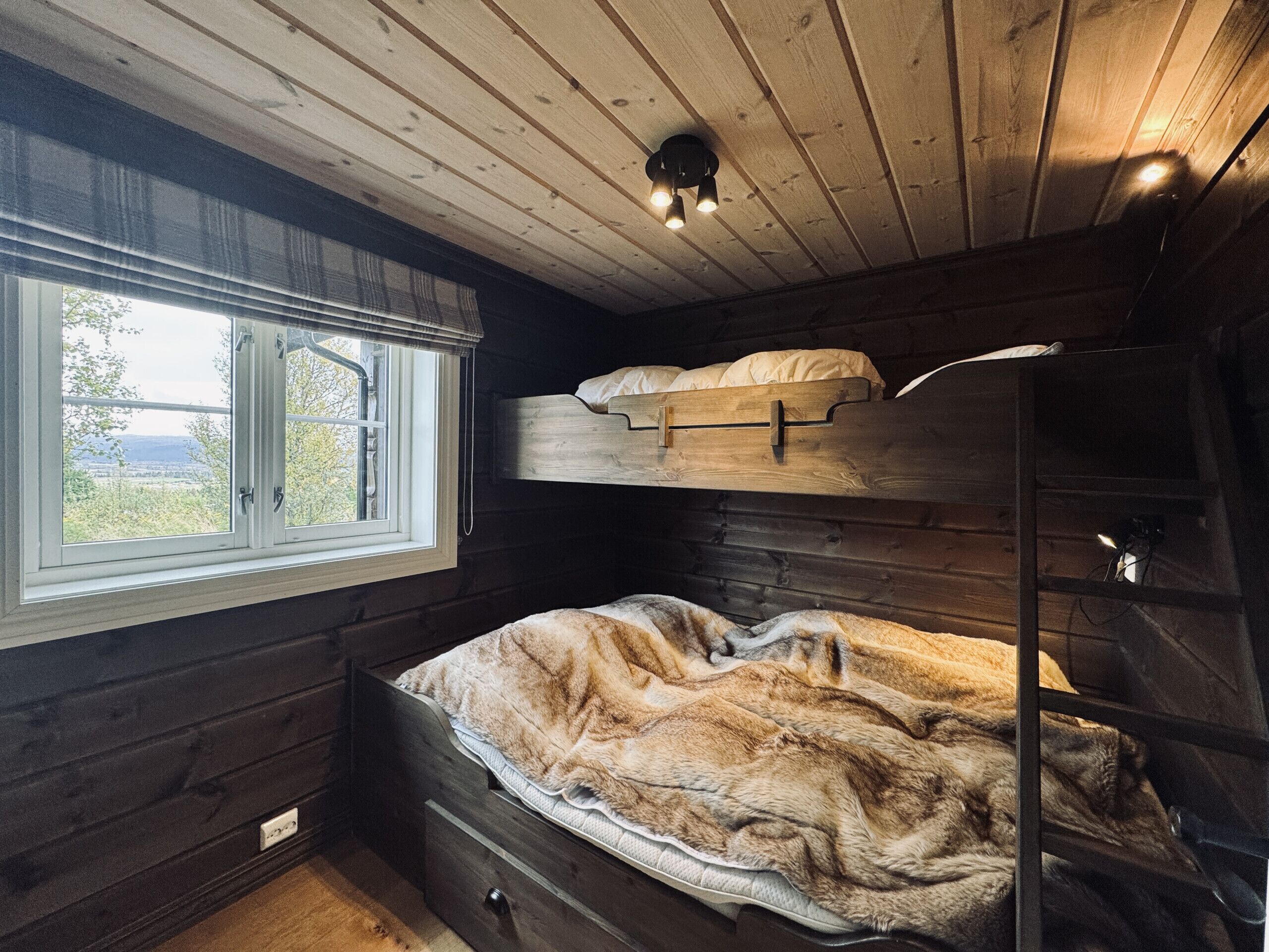 Uma cama beliche com janela, luminária no teto e piso de madeira.