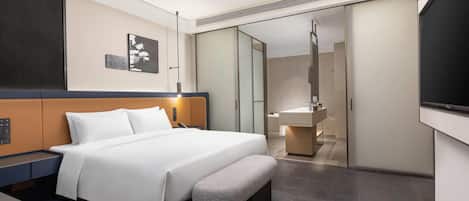A modern hotel room with a large bed, bedside table, bench, and a bathroom with a mirror and sink.