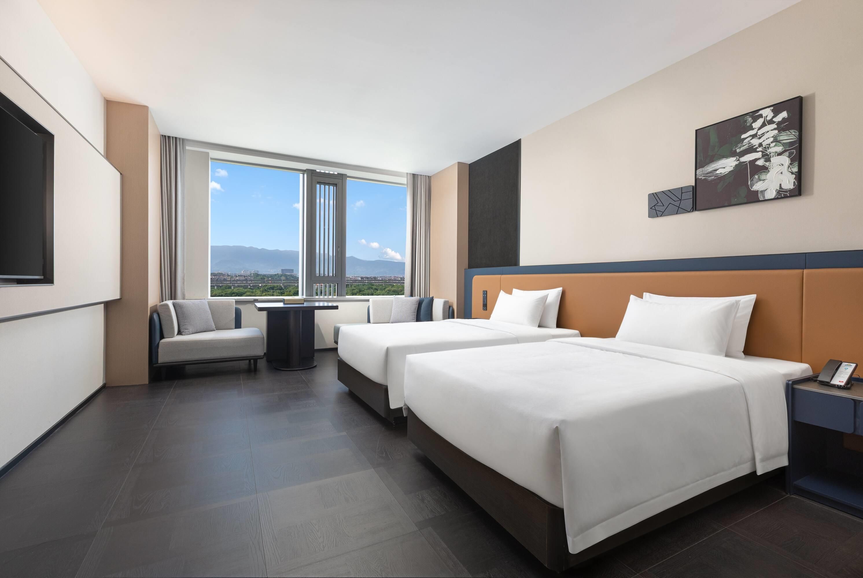 A hotel room with two beds, a sofa, a dining table, and a large window offering a city view.
