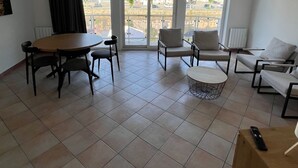 Ein großzügiger Wohnbereich mit Esstisch, Stühlen, einer Couch und einem Couchtisch. Große Fenster bieten Blick auf die Straße.