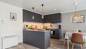 A modern kitchen with dark cabinets, a central island, pendant lighting, and a dining area with a table and chairs. - Valen Brygge, Digermulen (Vågan)