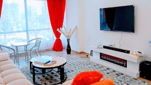A modern living room with a sofa, a round coffee table, a TV mounted on the wall, and a window with red curtains. - Marina Bay View Palace Apartment (Nairobi)