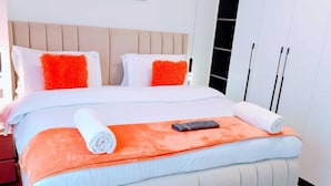 A neatly made bed with white and orange bedding, a remote control, and rolled towels on a room with a beige headboard and white walls. - Marina Bay View Palace Apartment (Nairobi)