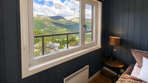 Een kamer met een groot raam en uitzicht op bergen, een tafeltje met een lamp, en een bed met kussens.