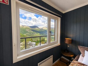 Habitación con amplia ventana que ofrece vista a la montaña, una mesita con lámpara y una cama con almohadas.