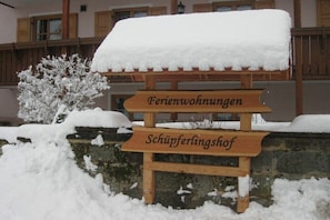 Ein Holzschild mit der Aufschrift 'Ferienwohnungen' und 'Schipferlingshof', vor verschneiter Landschaft und einem Gebäude im Hintergrund.
