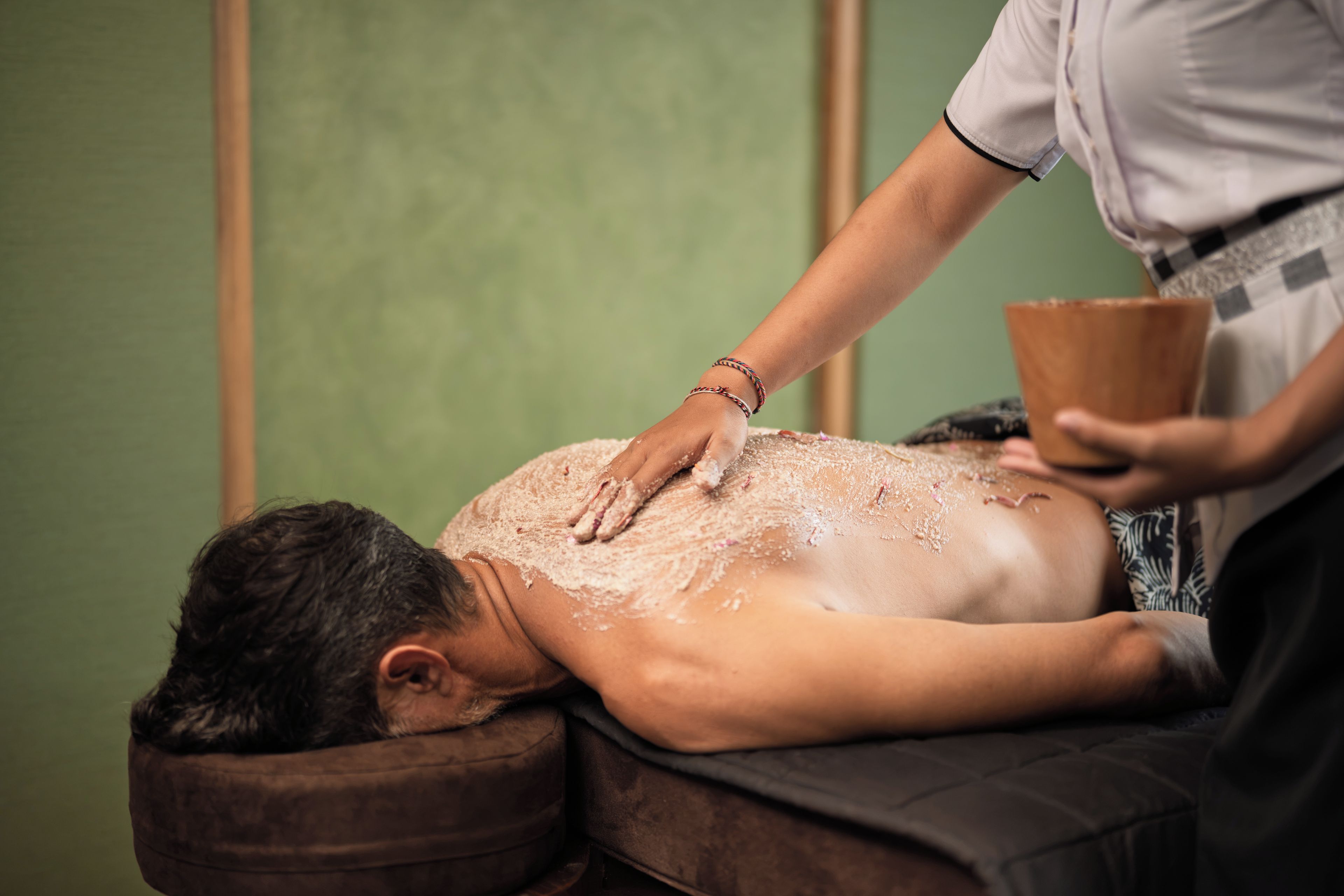 A person receiving a back massage in a room with a green wall and a wooden headrest.