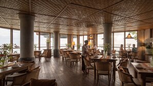 Un restaurante con amplios ventanales, mesas y sillas de madera, y un techo de paja.