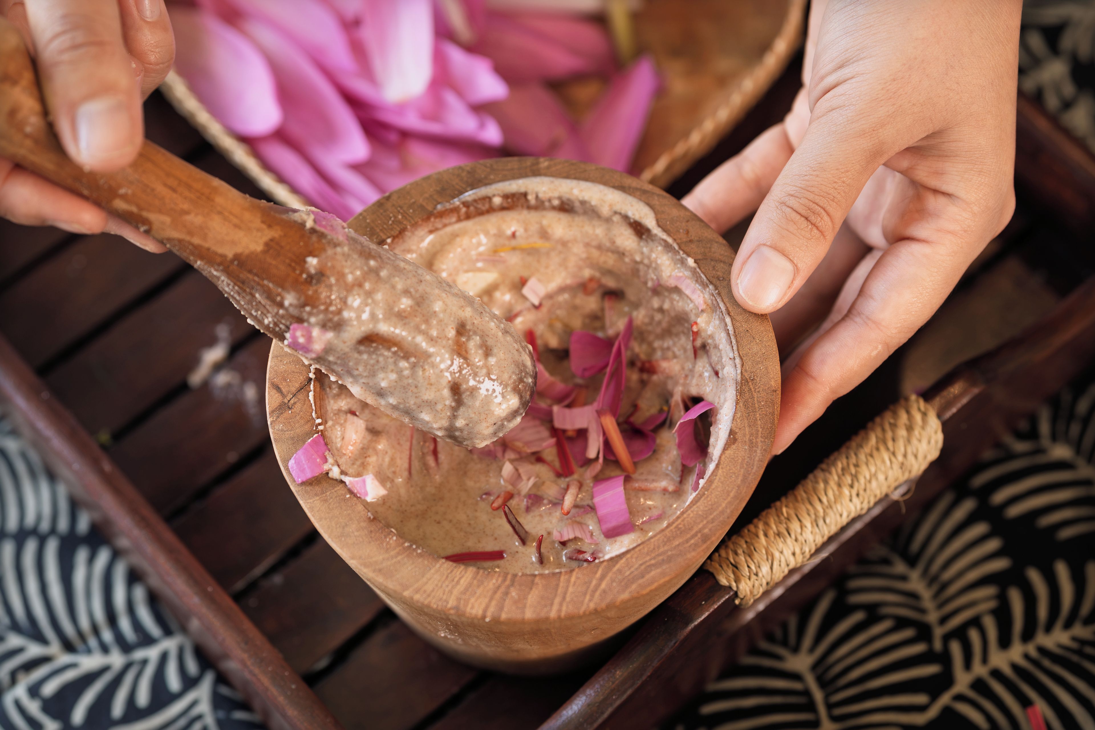 A wooden bowl with a spoon mixing a creamy substance, surrounded by pink petals.