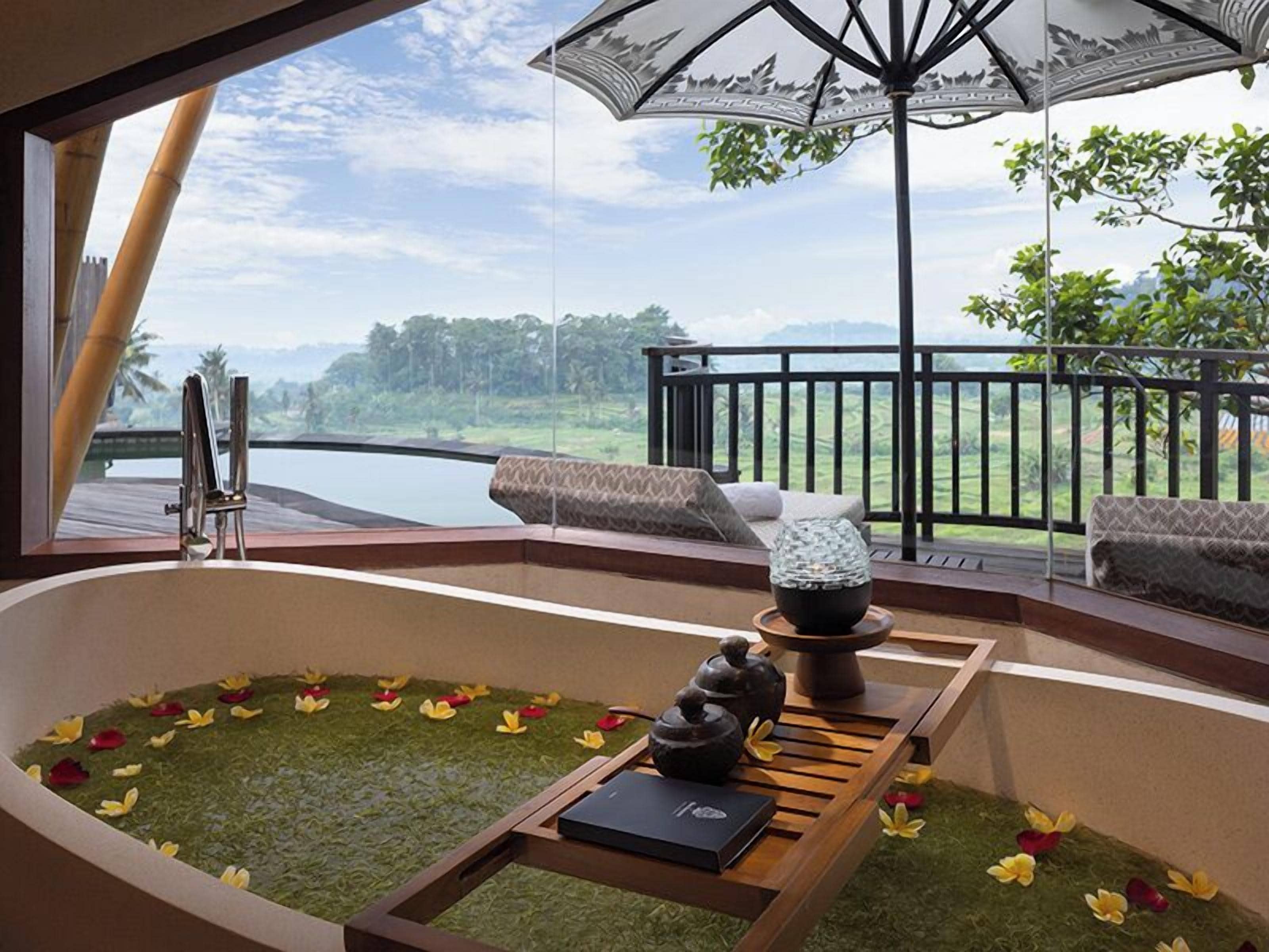 A room with a large bathtub, a table with a book, and a view of a lake and greenery.