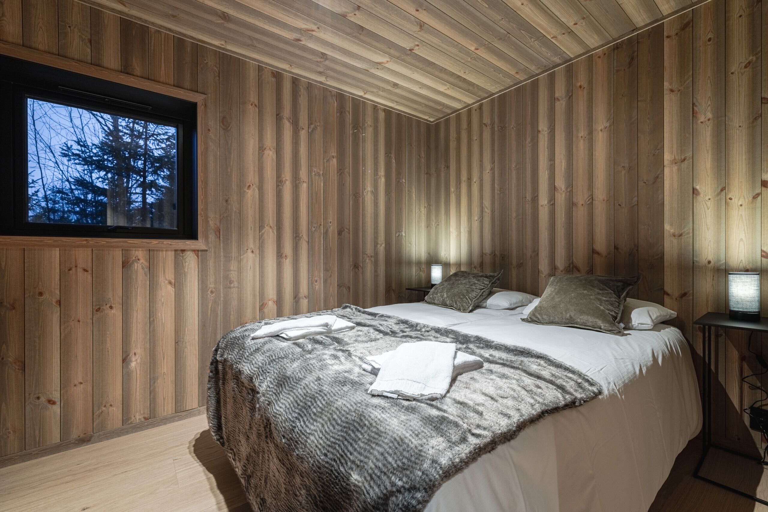Une chambre à coucher aux murs lambrissés, avec un lit, des oreillers, une couverture et une fenêtre donnant sur des arbres.