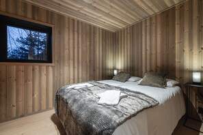 Une chambre à coucher aux murs lambrissés, avec un lit, des oreillers, une couverture et une fenêtre donnant sur des arbres.