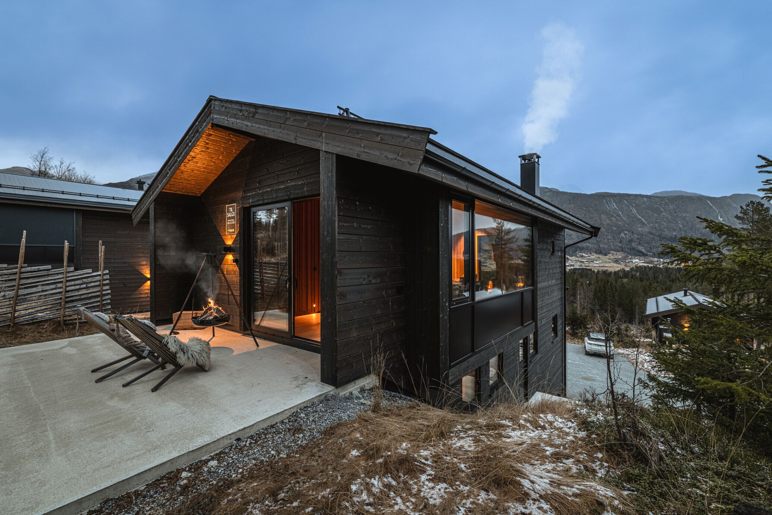 Une cabane moderne en bois, dotée d’une cheminée, de grandes fenêtres et offrant une vue sur le paysage environnant.