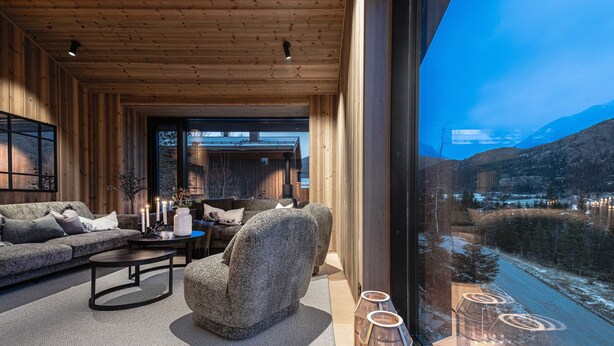 Un interno moderno con soffitto in legno, un divano, una poltrona, un tavolino da caffè e vista su un paesaggio innevato attraverso ampie finestre.