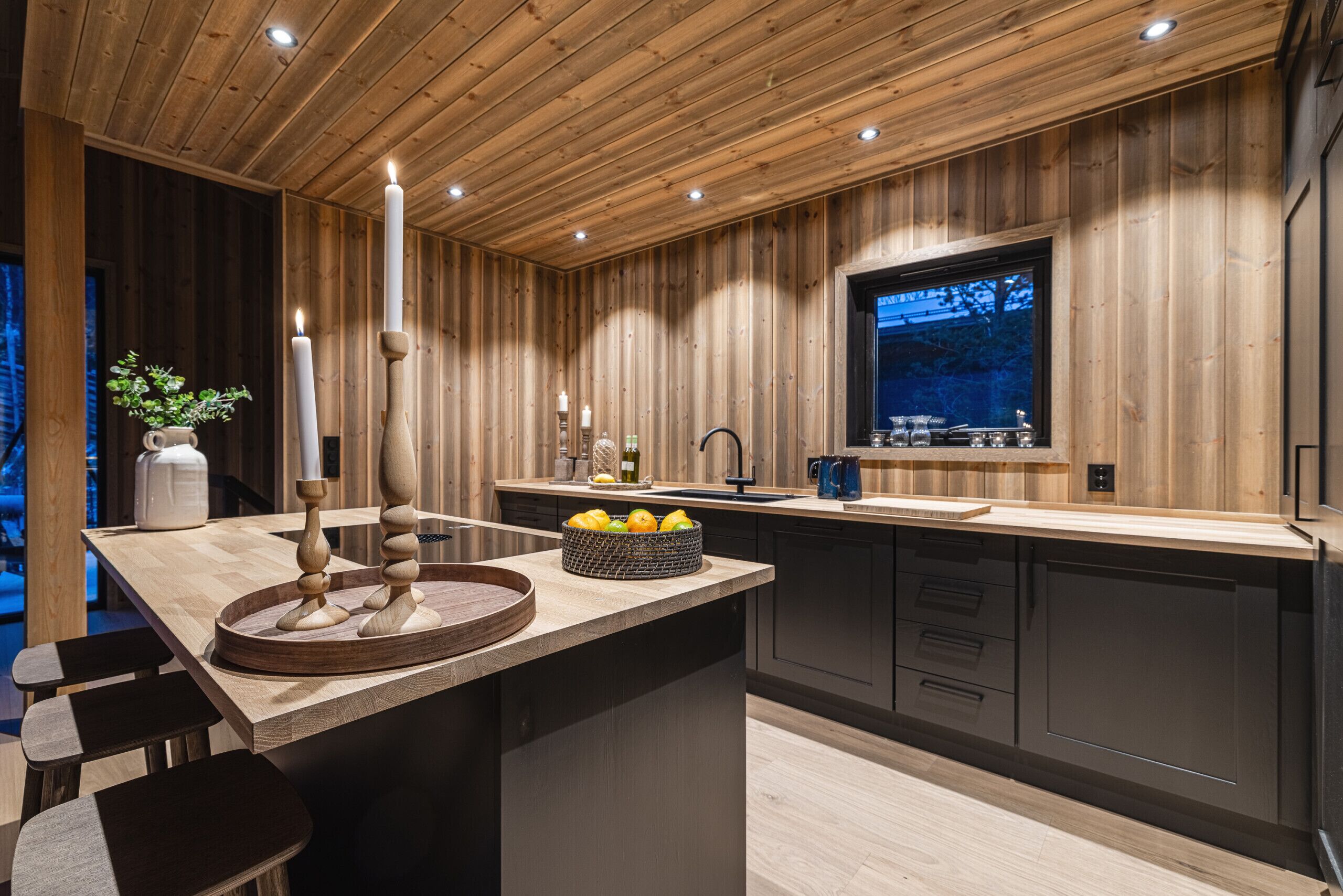 A modern kitchen with wooden paneling, a central island with a fruit bowl, and a window overlooking a forested area.