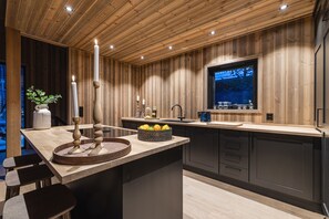 Une cuisine moderne avec un lambris en bois, un îlot central agrémenté d’un panier de fruits et une fenêtre donnant sur une zone boisée.