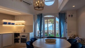 Een moderne keuken met inbouwapparatuur, een eethoek met een ronde tafel en blauwe stoelen, en uitzicht op zee door een groot raam.
