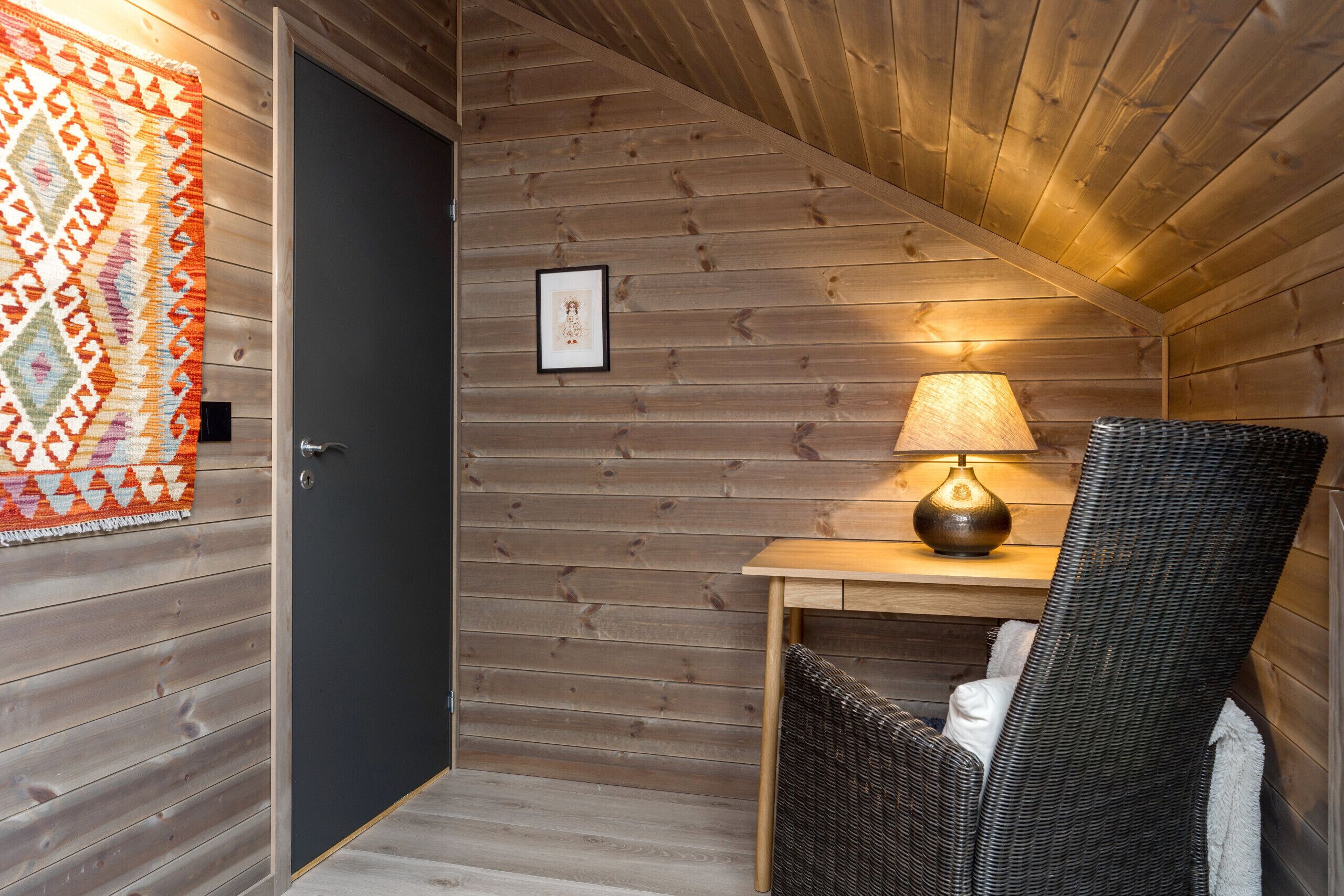 Interior de madera con una puerta, un cuadro enmarcado, una mesa con lámpara y una silla de mimbre.