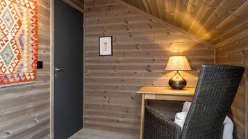 Interior de madera con una puerta, un cuadro enmarcado, una mesa con lámpara y una silla de mimbre.