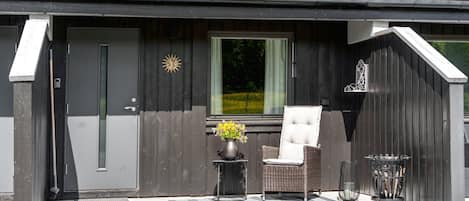 A modern building with a dark exterior, a front door, a window, a chair, a small table with a vase of flowers, and a metal fire pit.