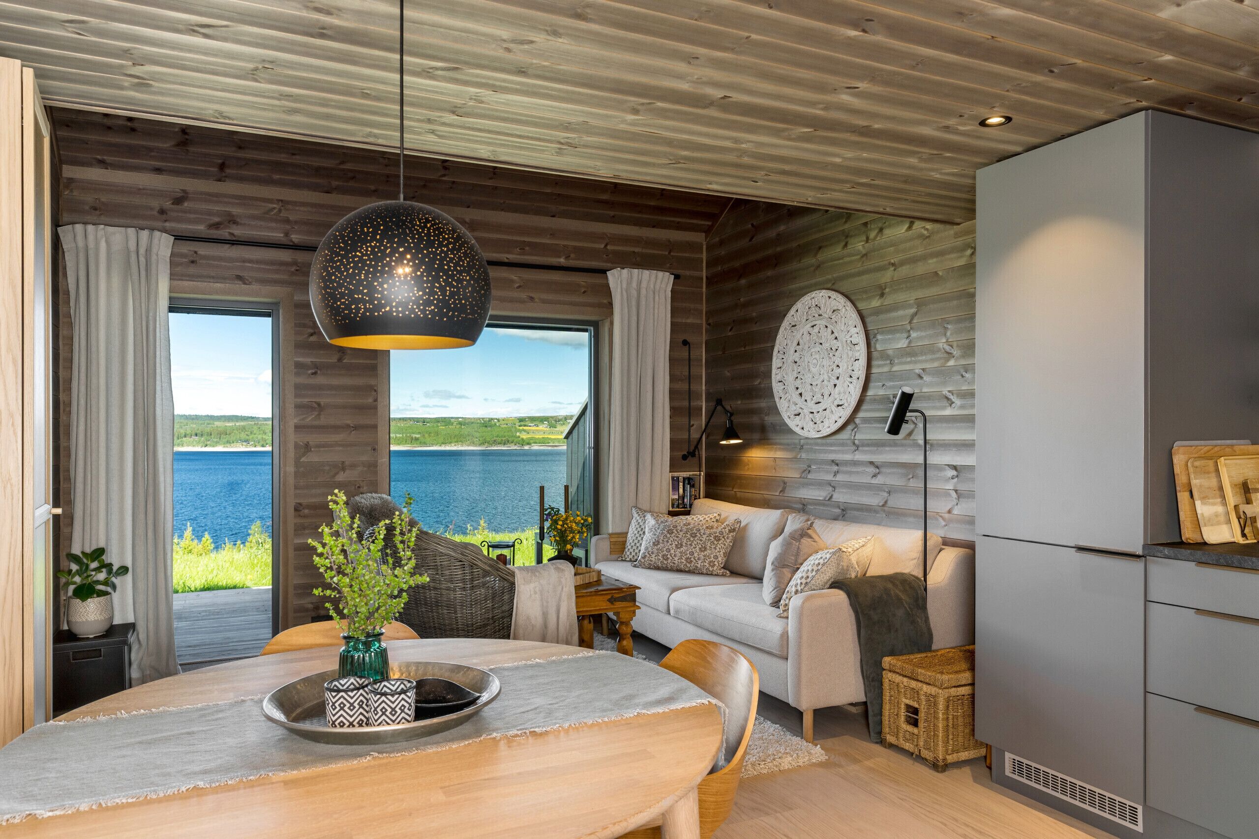 A modern interior with a dining area, sofa, and kitchen, featuring wooden walls and large windows overlooking a lake.
