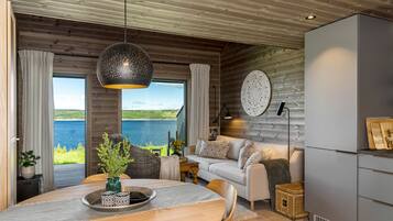 A modern interior with a dining area, sofa, and kitchen, featuring wooden walls and large windows overlooking a lake.