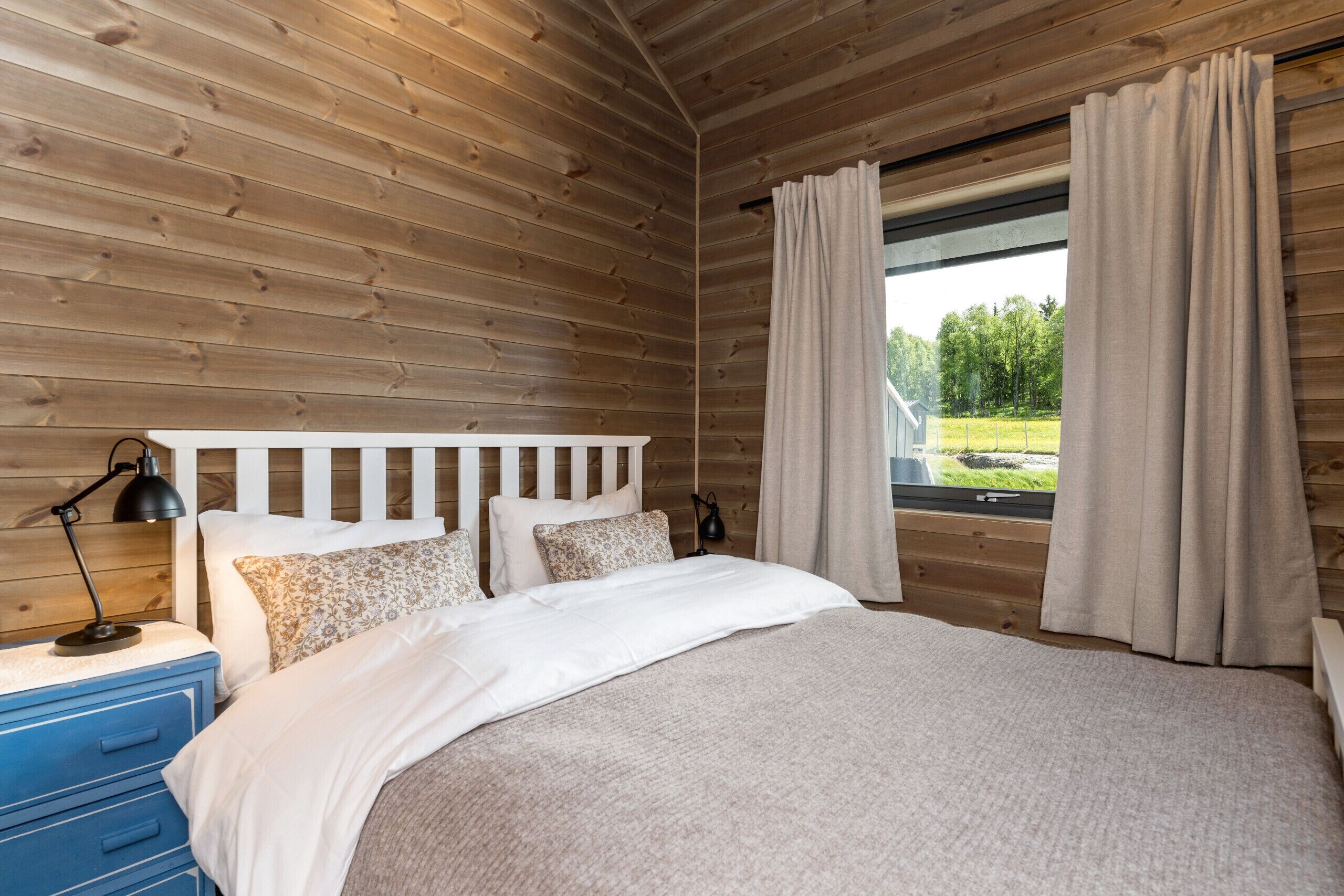Ein Zimmer mit Holzwänden, einem Bett, einem Nachttisch, einer Lampe und einem mit Vorhängen drapierten Fenster.
