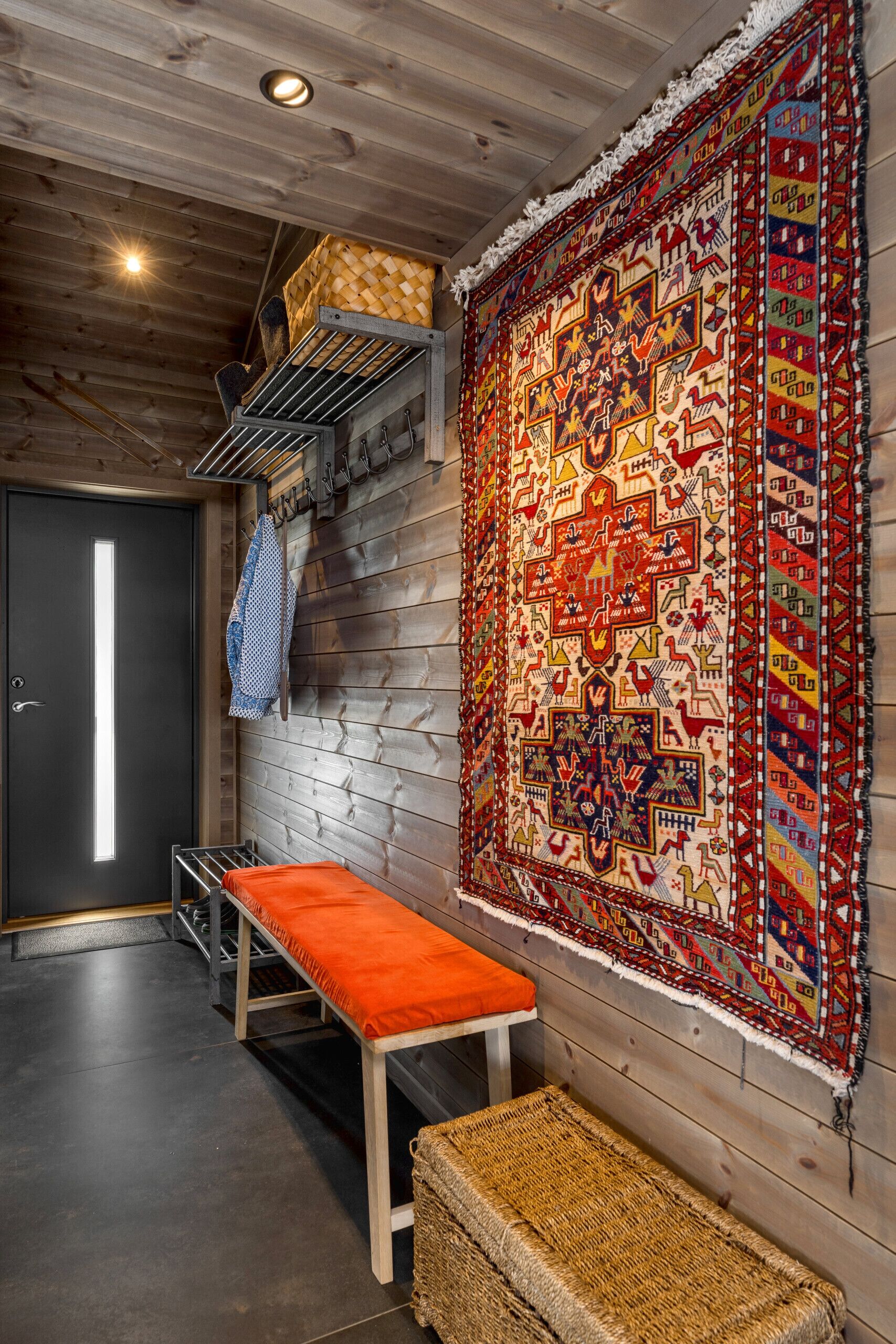 A room with a bench, a woven bench, a large patterned rug, and a door.