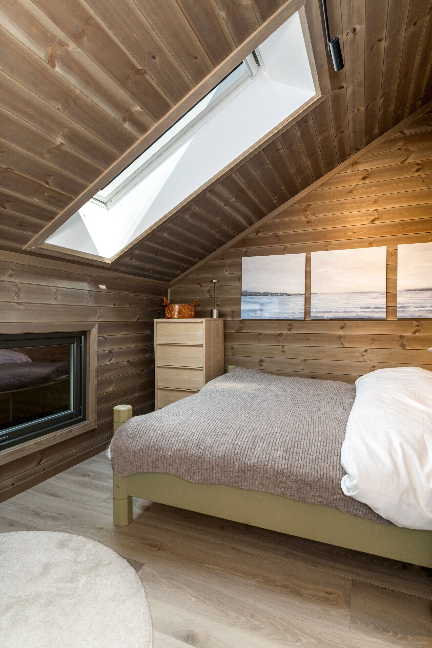 Ein Schlafzimmer mit Oberlicht, Holzwänden, einem Bett, einer Kommode und einem Fenster.