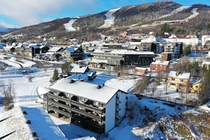 Ein mehrstöckiges Gebäude mit Balkonen, im Hintergrund ein Skigebiet und verschneiter Boden.