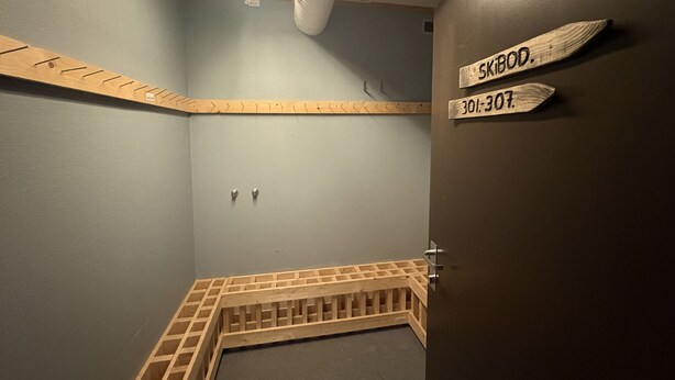 Une salle de bain avec un banc en bois, un porte-serviettes et une porte sur laquelle est apposée une pancarte indiquant « SKIDDO » et un numéro de téléphone.