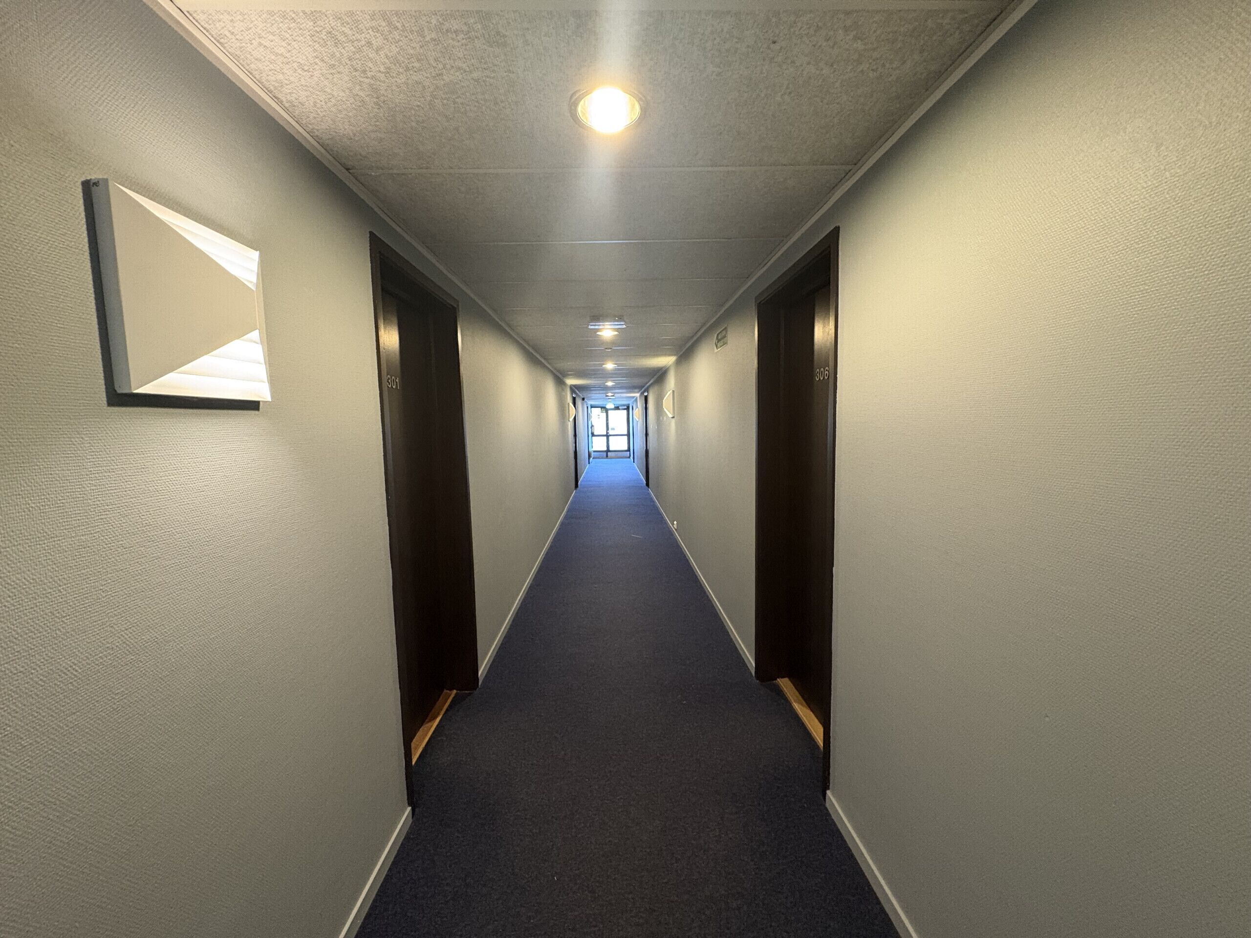 Un couloir d’hôtel avec des planchers recouverts de moquette, des portes de chaque côté et une lumière au bout.