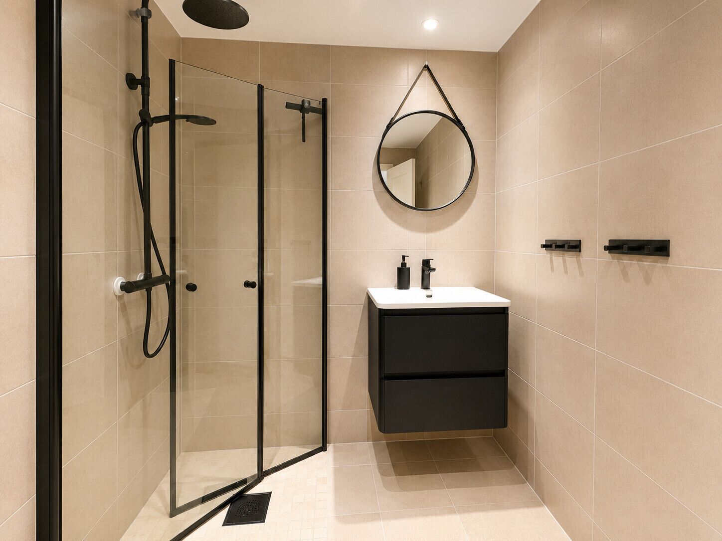 Un baño moderno con cabina de ducha de vidrio, un espejo redondo, un mueble de tocador negro con encimera blanca y paredes con azulejos color beige.