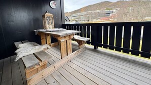 Une terrasse en bois avec une table et des bancs, une lanterne, et une vue sur un paysage vallonné parsemé de bâtiments.